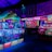 An indoor arcade filled with vibrant gaming machines lit with neon blue and pink lights. The scene includes various arcade games with flashing screens and colorful displays, and a large overhead structure resembling a green snake. The background shows illuminated signs and themed decor, contributing to an immersive entertainment atmosphere.