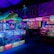 An indoor arcade filled with vibrant gaming machines lit with neon blue and pink lights. The scene includes various arcade games with flashing screens and colorful displays, and a large overhead structure resembling a green snake. The background shows illuminated signs and themed decor, contributing to an immersive entertainment atmosphere.