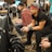 Several people are sitting in a room with gaming consoles and monitors, focusing intently on their screens. The setting appears to be a casual gaming event, with participants holding controllers while playing video games. The room has a casual atmosphere with chairs and tables set up for gaming.