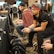 Several people are sitting in a room with gaming consoles and monitors, focusing intently on their screens. The setting appears to be a casual gaming event, with participants holding controllers while playing video games. The room has a casual atmosphere with chairs and tables set up for gaming.