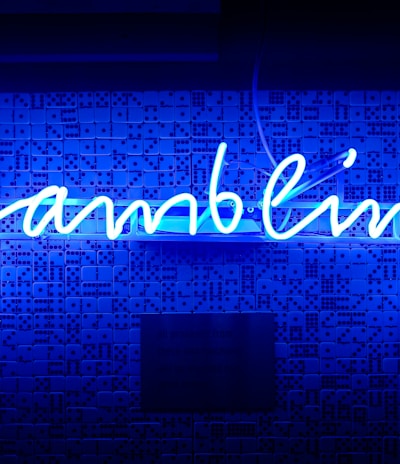 A bright blue neon sign spells out the word 'gambling' in cursive script. The background features a textured wall covered in domino tiles, also illuminated by the blue light, creating a vivid and visually striking scene.