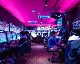 A vibrant arcade with rows of gaming machines adorned with colorful screens. Several people are immersed in playing games, seated on stools in a dimly lit room accented by an intense pink ceiling light. The atmosphere is lively and engaging.