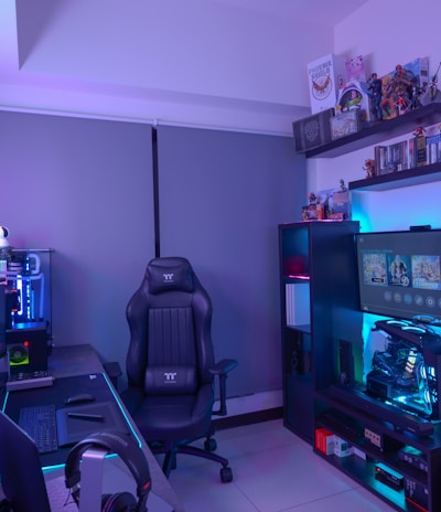 A modern gaming setup featuring a sleek gaming chair, dual monitors on a desk, and LED lighting. A gaming console and various decorations are displayed on shelves, creating an organized and vibrant gaming environment.