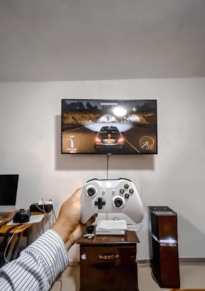 A person is holding a gaming controller in front of a flat-screen television displaying a racing video game. The room has a computer setup on a wooden desk with cables, a desktop computer monitor, and other accessories. The focus is on the interaction between the person and the gaming environment.
