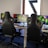A row of gaming stations equipped with large monitors, gaming chairs, keyboards, and headphones. The setup is organized and ready for gaming, featuring a colorful screen display. The room has a modern and tech-savvy ambiance.