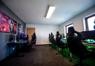 A well-lit gaming room features multiple gaming stations with high-backed chairs and monitors. The room has a minimalist setup with green and black color tones. Large posters adorn the walls, and the room appears clean and organized.