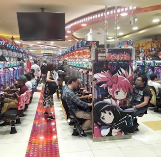 A bustling arcade filled with people playing various gaming machines. Brightly colored displays and animated characters are prominent on posters and screens. The atmosphere is lively with patrons seated in rows, focusing on the games. Overhead signage and electronic scoreboards are visible, contributing to the busy and vibrant environment.