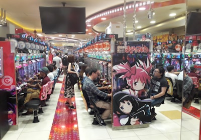 A bustling arcade filled with people playing various gaming machines. Brightly colored displays and animated characters are prominent on posters and screens. The atmosphere is lively with patrons seated in rows, focusing on the games. Overhead signage and electronic scoreboards are visible, contributing to the busy and vibrant environment.