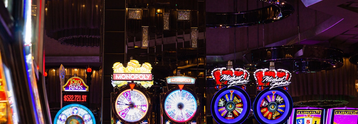 A lively casino scene featuring a row of bright, colorful slot machines with themes like Monopoly, Michael Jackson, and Lord of the Rings. The gaming machines display a variety of flashing lights and digital screens, creating an energetic and exciting atmosphere. Reflections of the lights are visible in glossy surfaces above the machines.