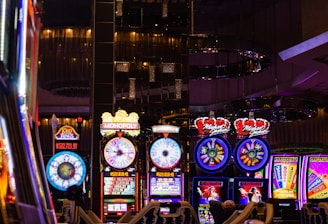 A lively casino scene featuring a row of bright, colorful slot machines with themes like Monopoly, Michael Jackson, and Lord of the Rings. The gaming machines display a variety of flashing lights and digital screens, creating an energetic and exciting atmosphere. Reflections of the lights are visible in glossy surfaces above the machines.