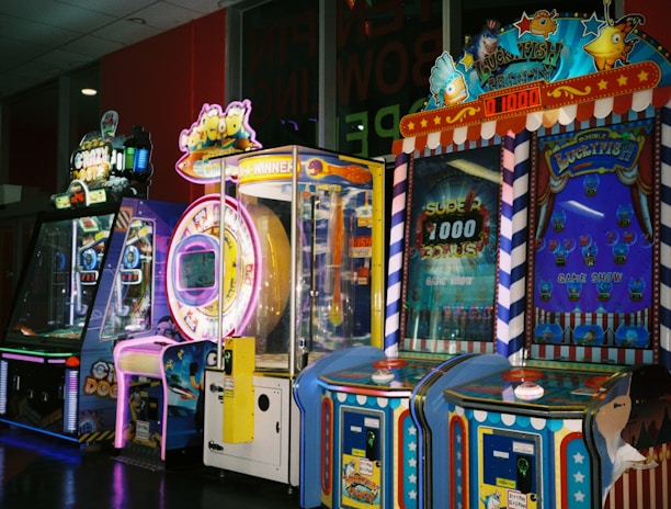 A row of brightly colored arcade game machines is arranged in a line against the wall. Each machine features vibrant lights and designs, with themes ranging from claw games to game show-inspired setups. The area has a playful and inviting atmosphere, suitable for entertainment or amusement centers.