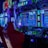 A vibrant arcade environment with several illuminated gaming machines. The image features arcade racing games with visible steering wheels, colorful control panels, and screens displaying game instructions. Bright LED lights in various colors illuminate the machines and surrounding area.