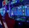 A vibrant arcade environment with several illuminated gaming machines. The image features arcade racing games with visible steering wheels, colorful control panels, and screens displaying game instructions. Bright LED lights in various colors illuminate the machines and surrounding area.